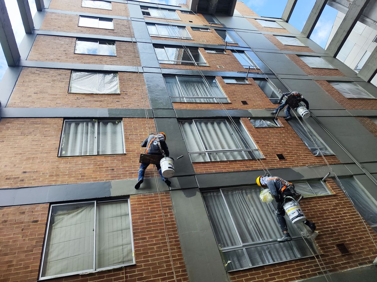 Tres operarios de Grupo Serviandina descolgados con arnés pintando la fachada de un edificio de ladrillo